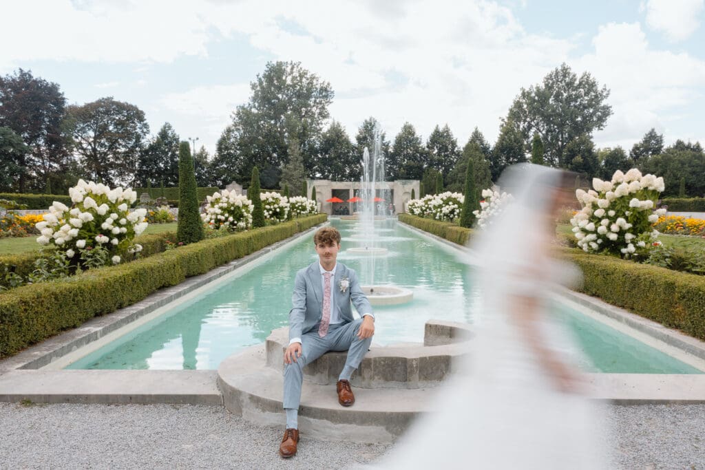 Groom seated beside the reflecting pool with the bride moving through the foreground
